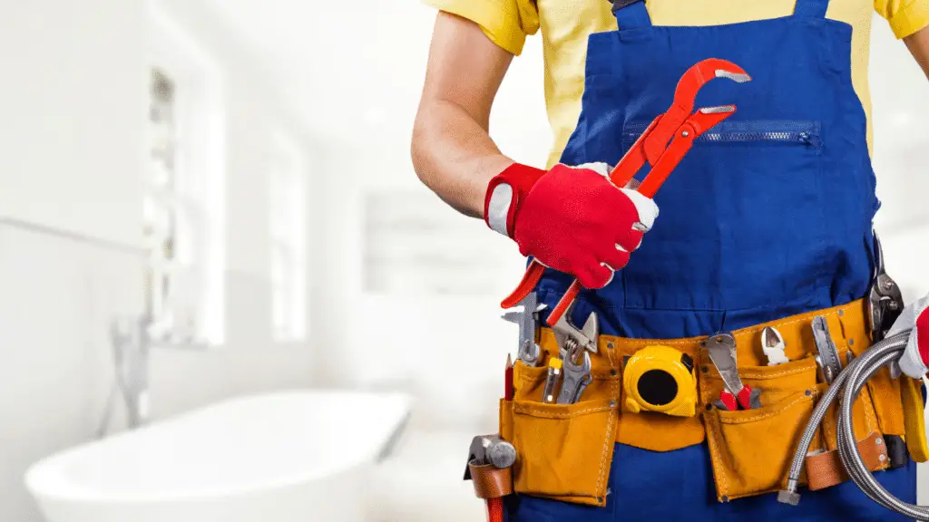 A man wearing a tool belt and holding a red wrench stands in a bathroom.