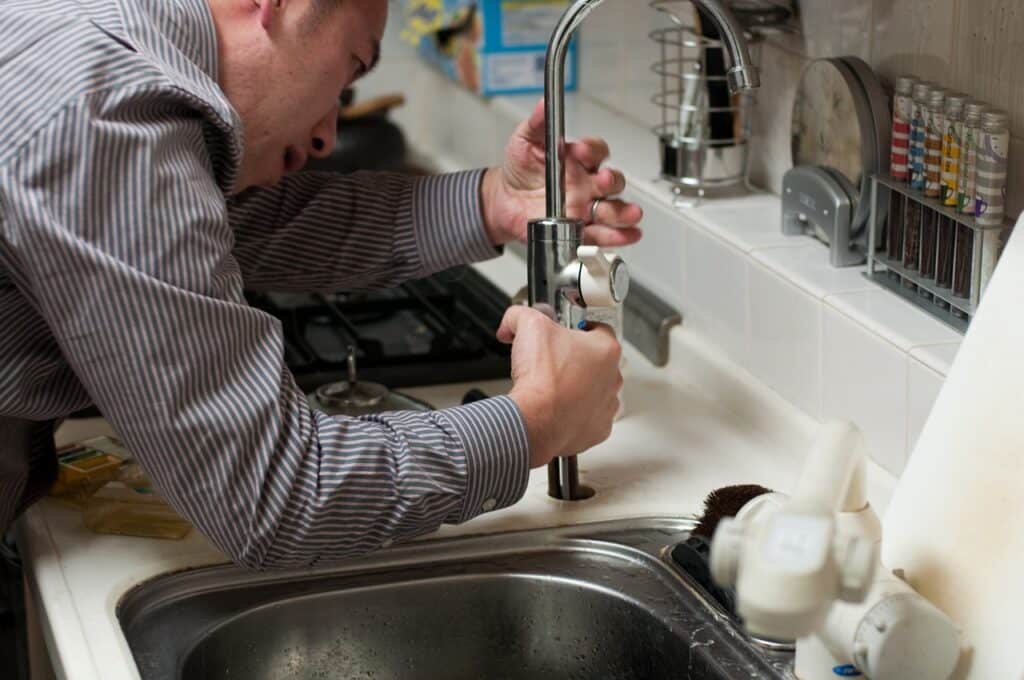 A man kneels beside a sink, diligently repairing plumbing with tools in hand, focused on the task at hand.