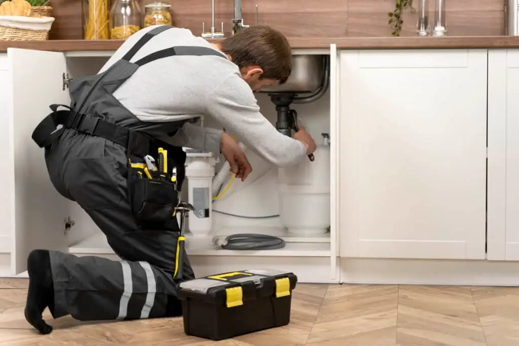 A plumber wearing overalls is kneeling and working on pipes under a kitchen sink.