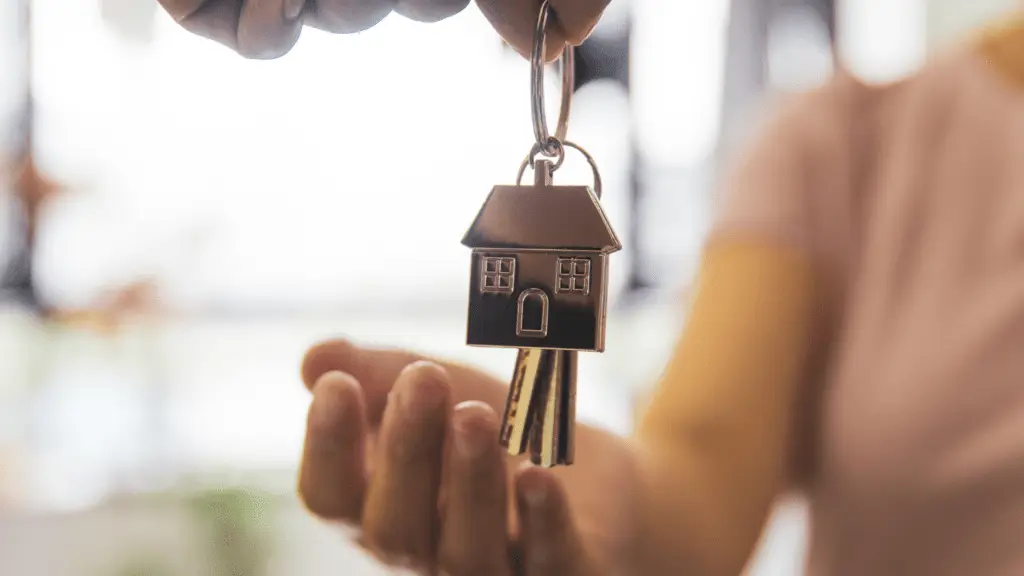 A house key with a house shaped keychain is handed from one person to another.